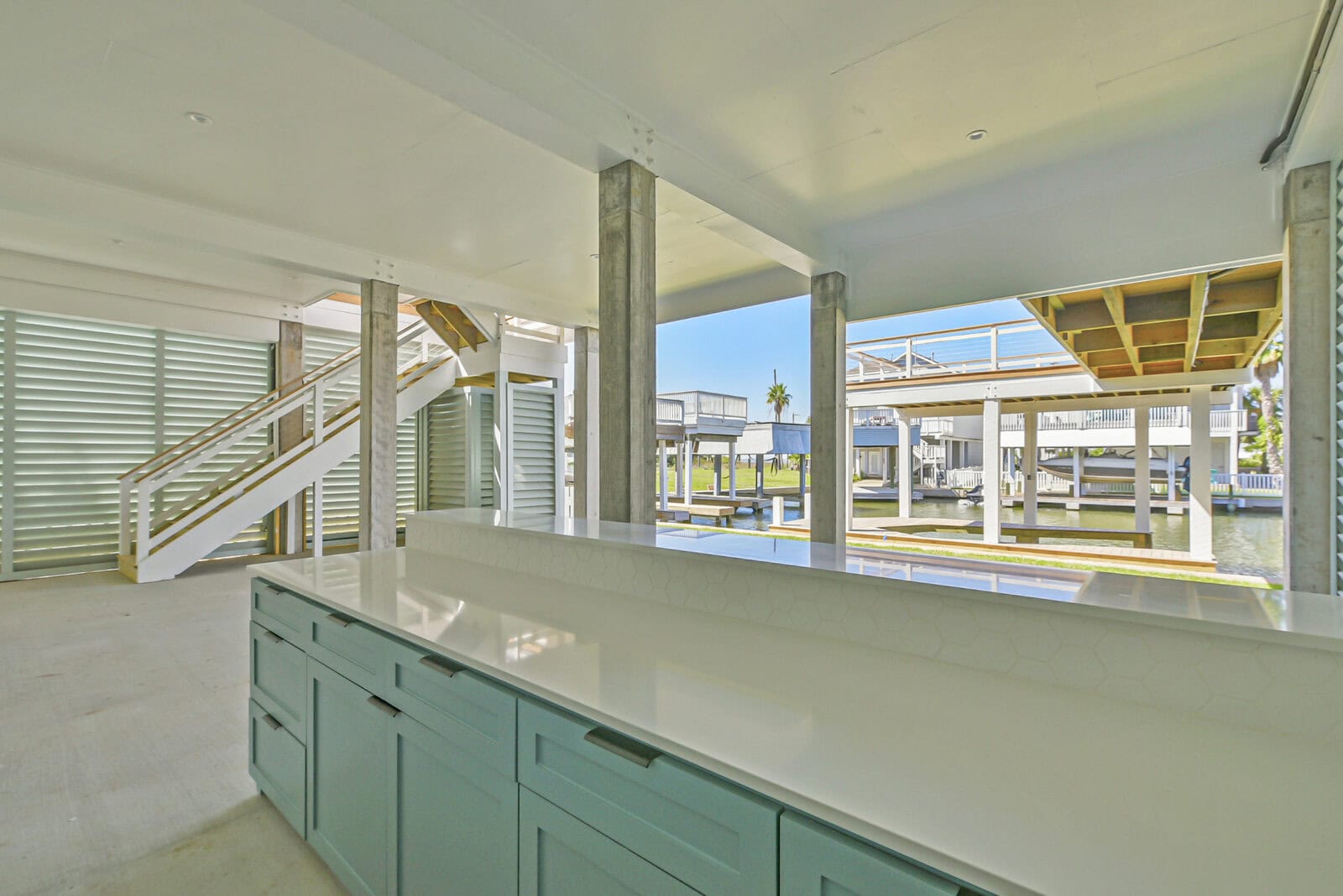 Open ground level beneath an elevated coastal home showing the concrete support columns, staircase and outdoor bar overlooking the canal