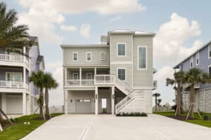 Front elevation of a completed pale green elevated coastal home built by Kai Custom Homes, raised above a ground-level garage with palm trees flanking the concrete driveway