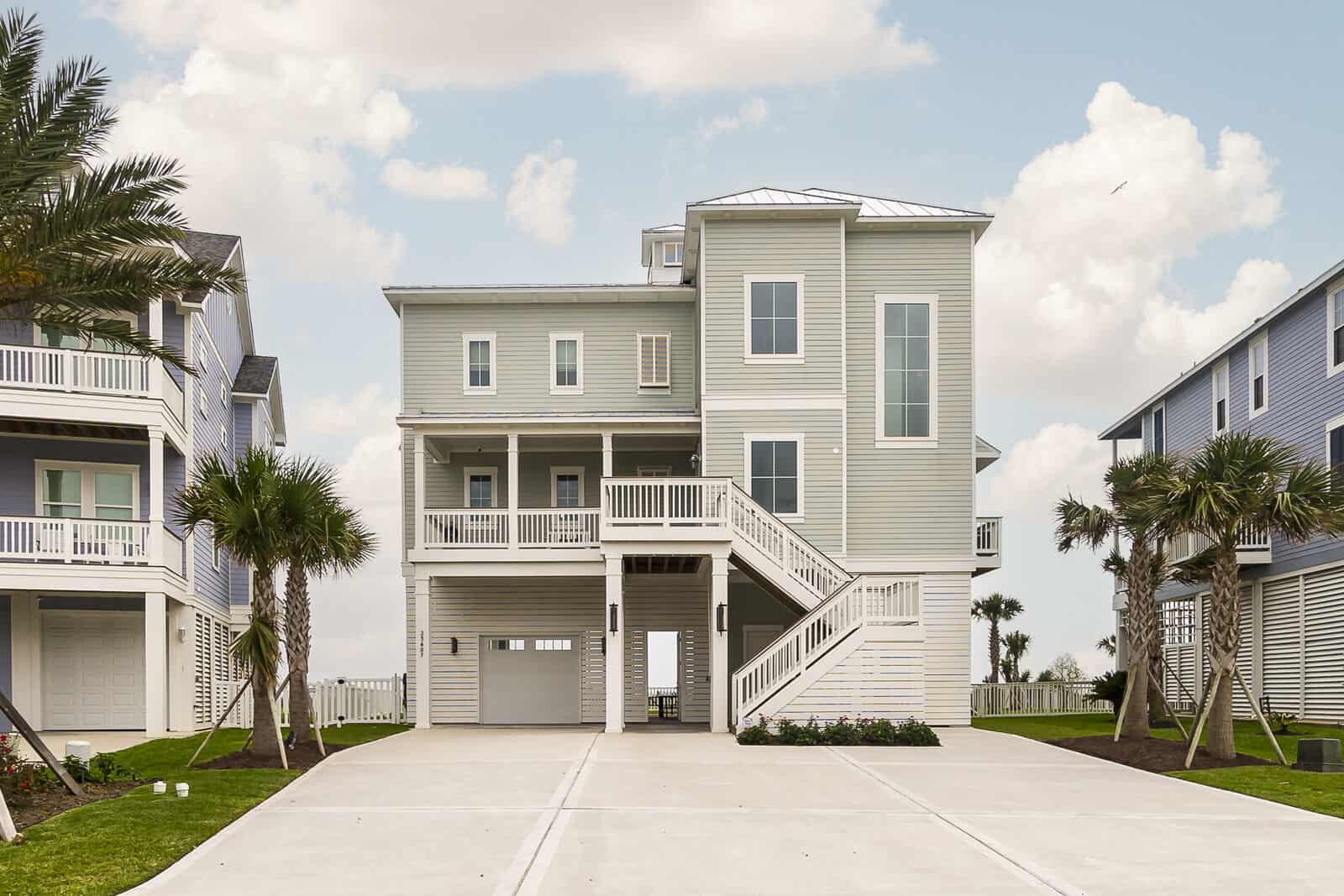Front elevation of a completed pale green elevated coastal home built by Kai Custom Homes, raised above a ground-level garage with palm trees flanking the concrete driveway