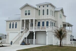Completed cream-colored three-story elevated coastal home by Kai Custom Homes, raised on columns above a ground-level garage with exterior stairs to the main living floor