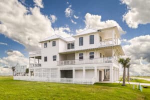 Homepage Side elevation of a completed white elevated coastal home by Kai Custom Homes showing stacked wraparound porches, metal roof and louvered ground-level enclosure beneath the raised structure