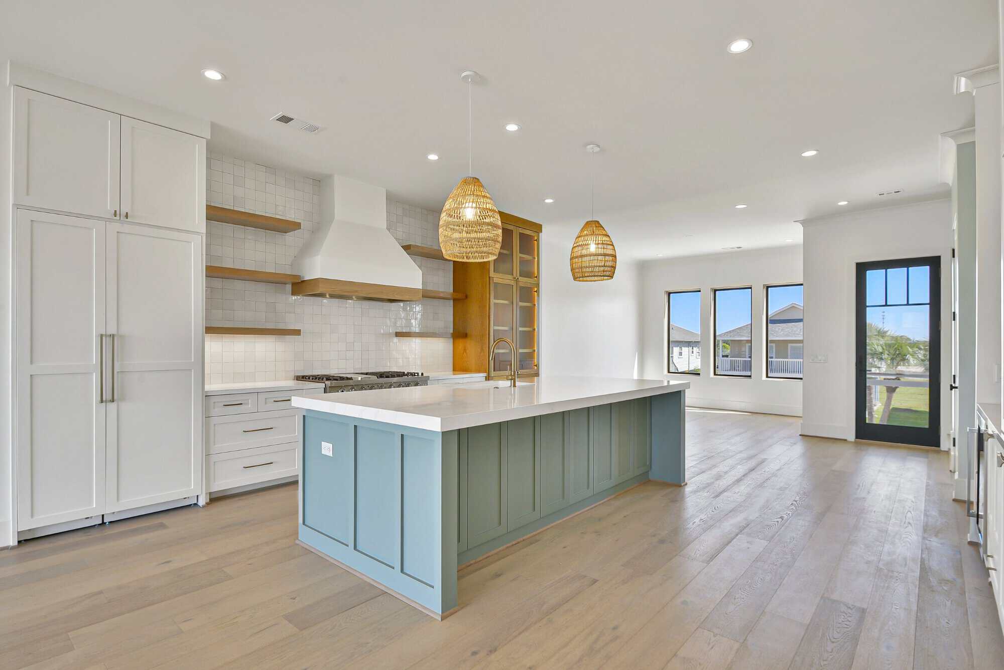 Open kitchen with a blue-gray island, rattan pendants and white-oak floors in a Kai Custom Homes build in Pirates Cove, Galveston