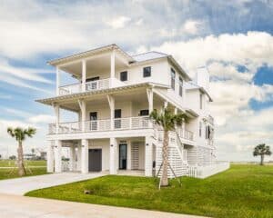 Homepage White three-story elevated coastal home with wraparound porches and a standing-seam metal roof, built by Kai Custom Homes in Sweetwater Cove on the Galveston West End